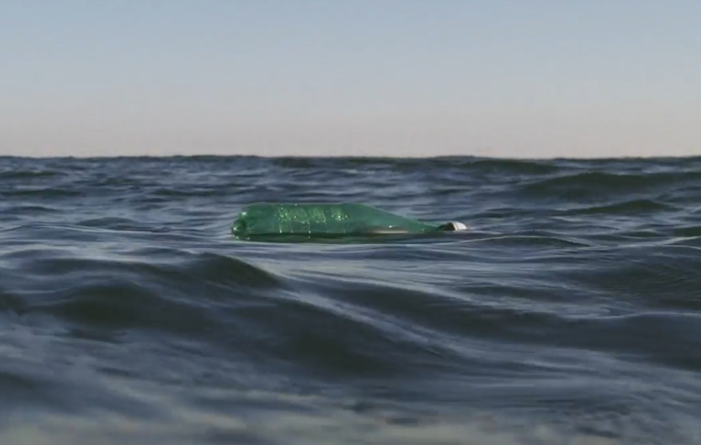 A green plastic bottle floats in the open ocean under a blue sky.