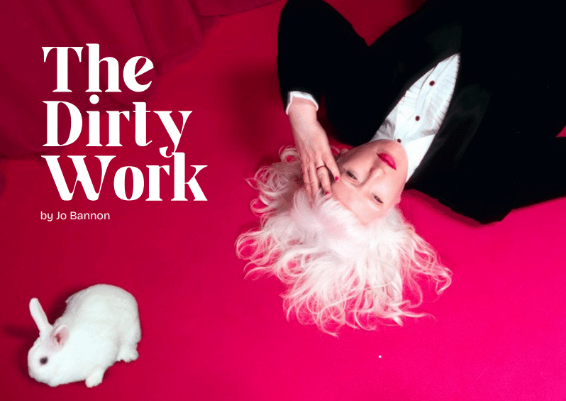 A woman in a tuxedo lies upon a hot pink carpet. Her white hair splays out over the floor. A white rabbit sits next to her. Text reads The Dirty Work by Jo Bannon.