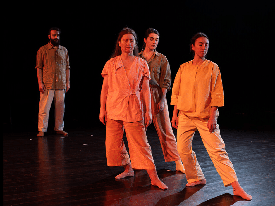 Three dancers stand in orange light, one leg extended along the floor. A fourth figure stand behind them on the left watching.